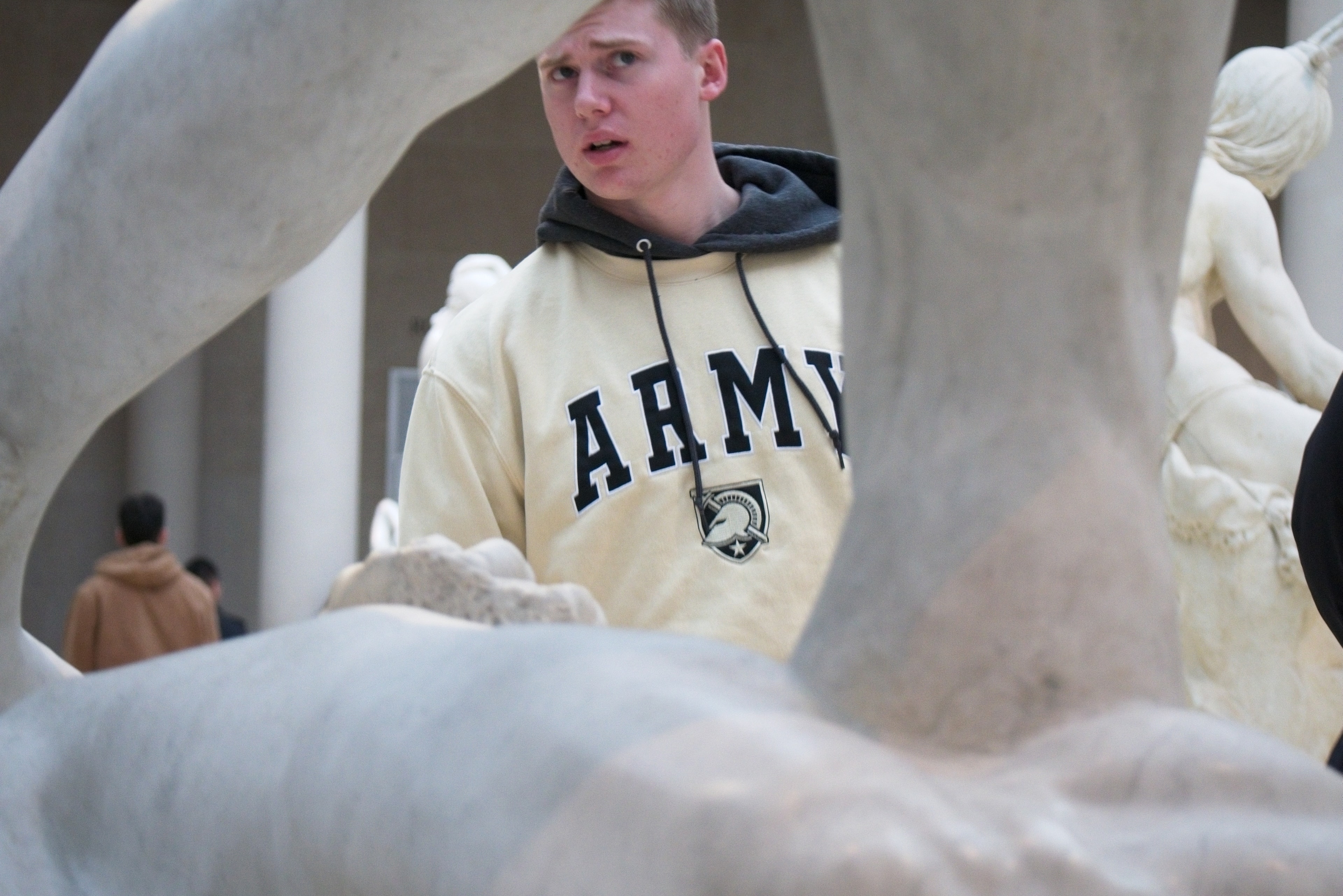 Young man in an Army hoodie peers through the limbs of a white marble sculpture in a museum courtyard