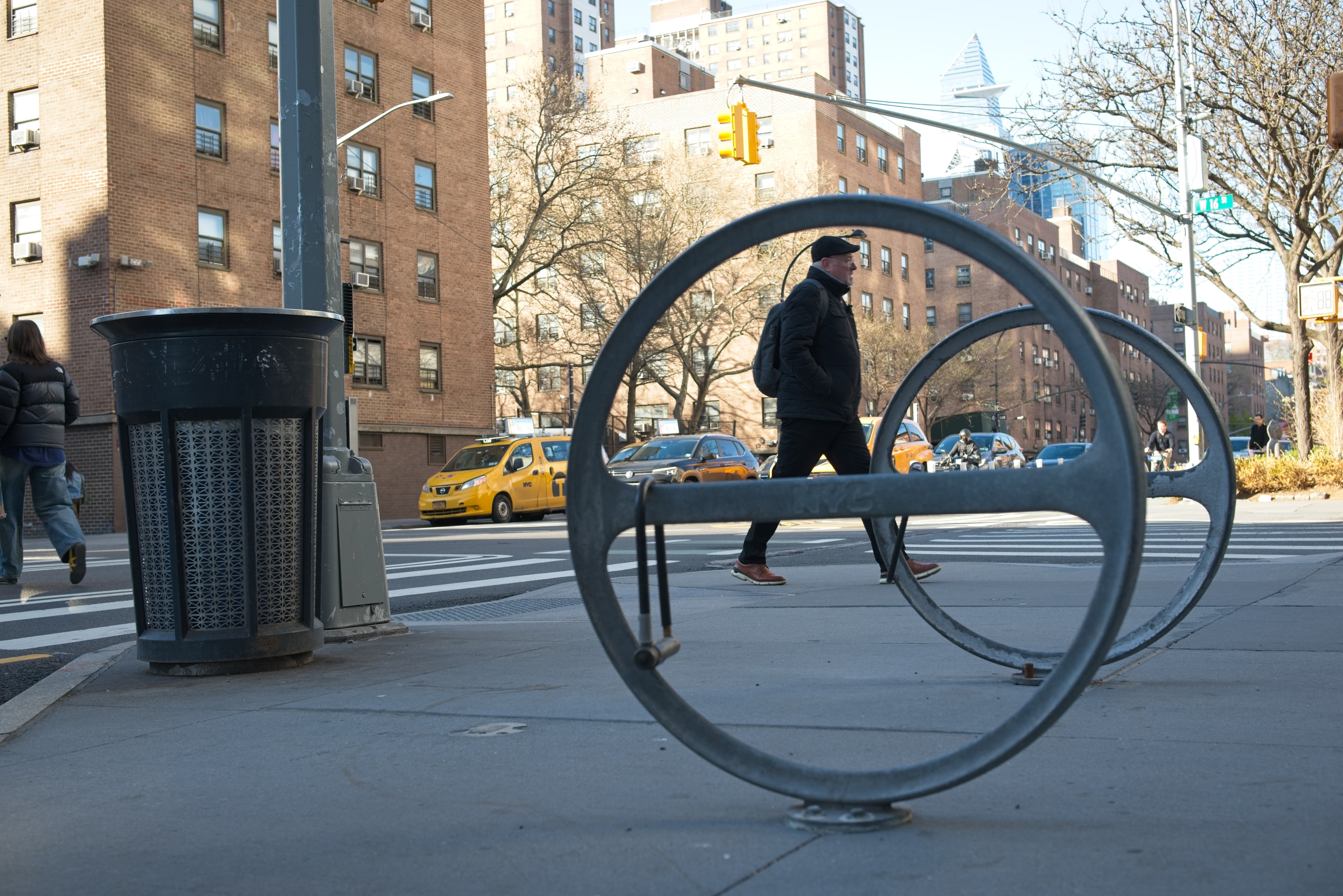 A circular metal bike rack frames a pedestrian crossing a busy NYC intersection with yellow taxis in the background