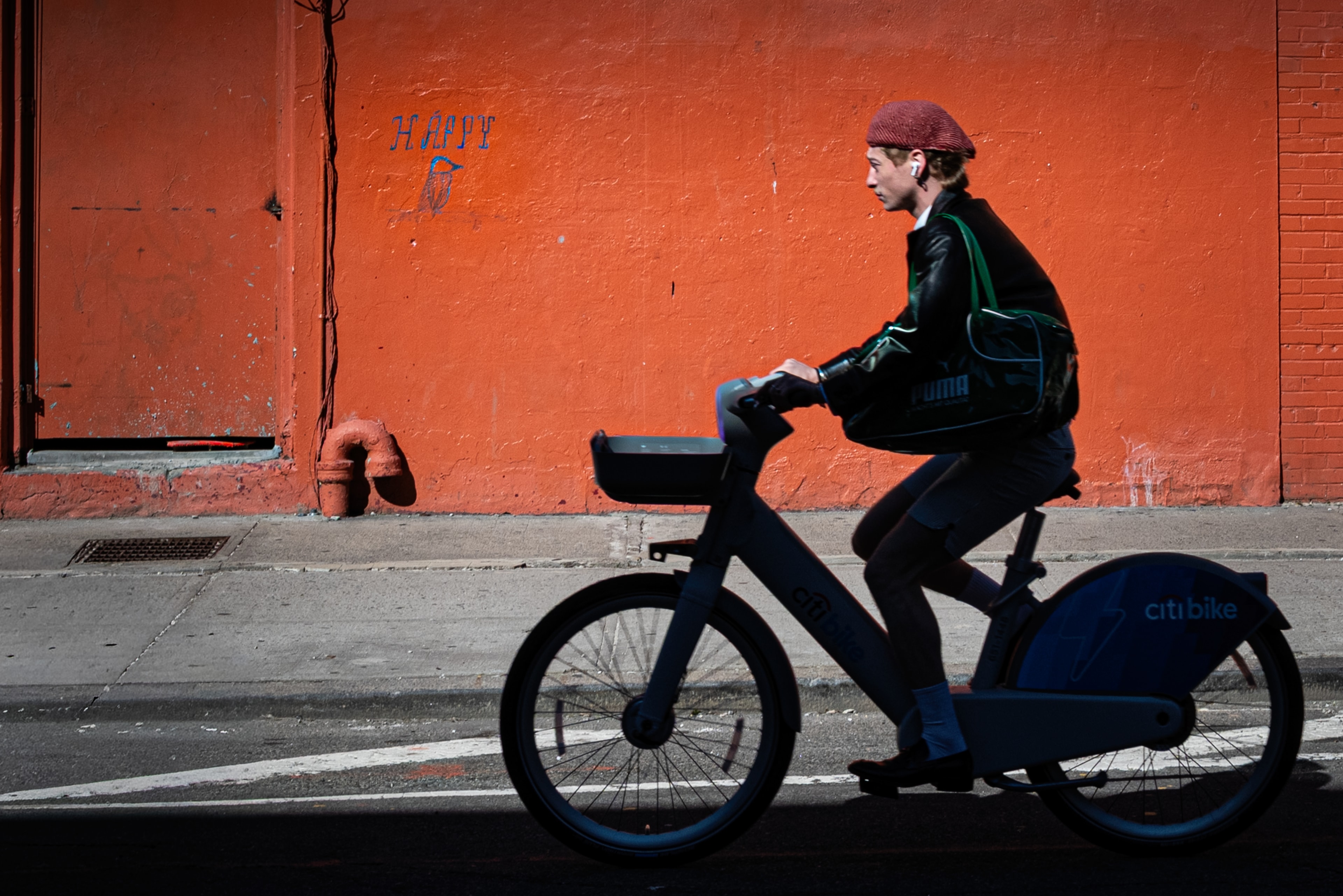 Cyclist on a Citi Bike passes in front of a vivid orange wall with 'HAPPY' graffiti
