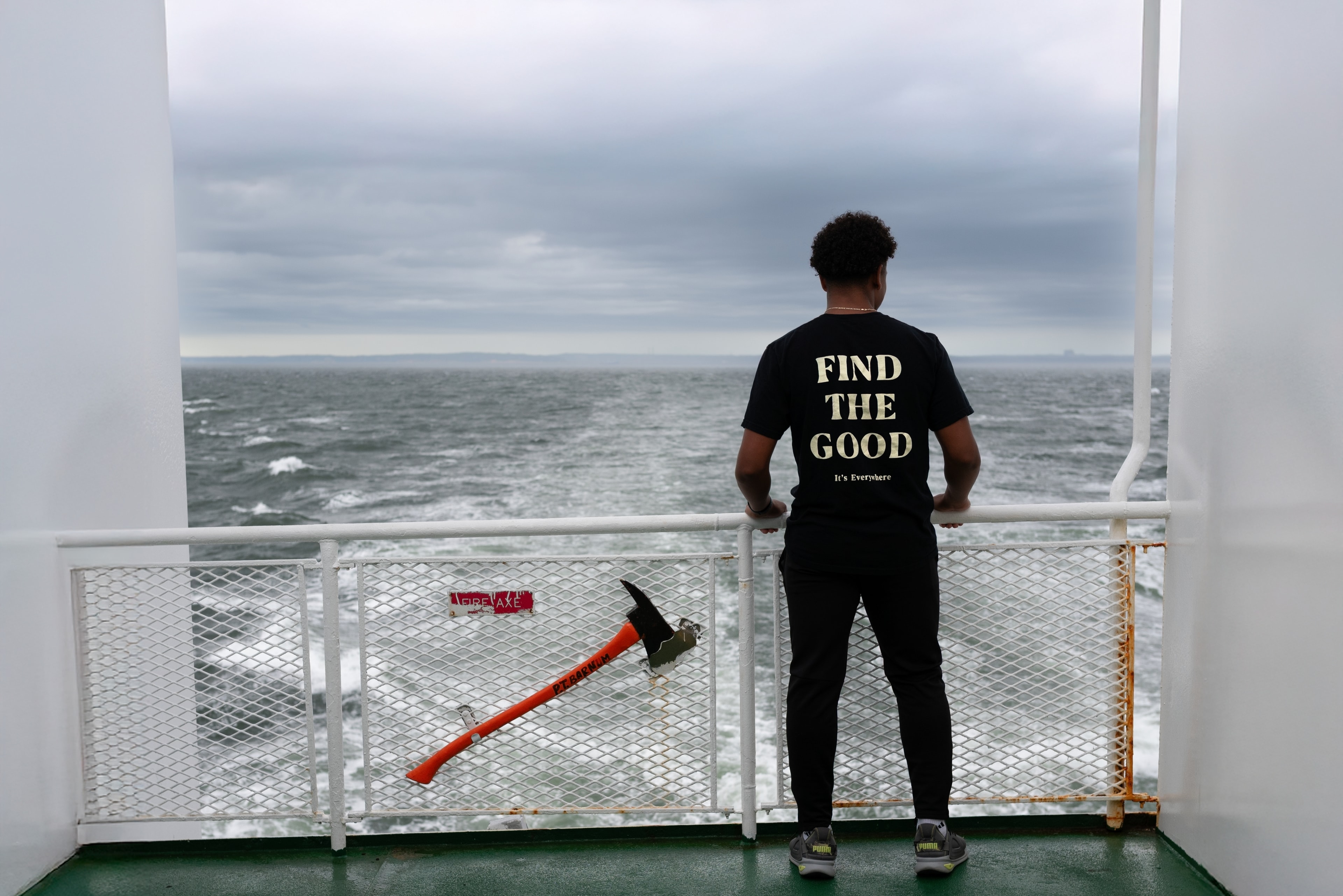 Young man in a 'Find The Good' t-shirt stands at a ferry railing gazing out at a stormy sea, a fire axe visible beside him