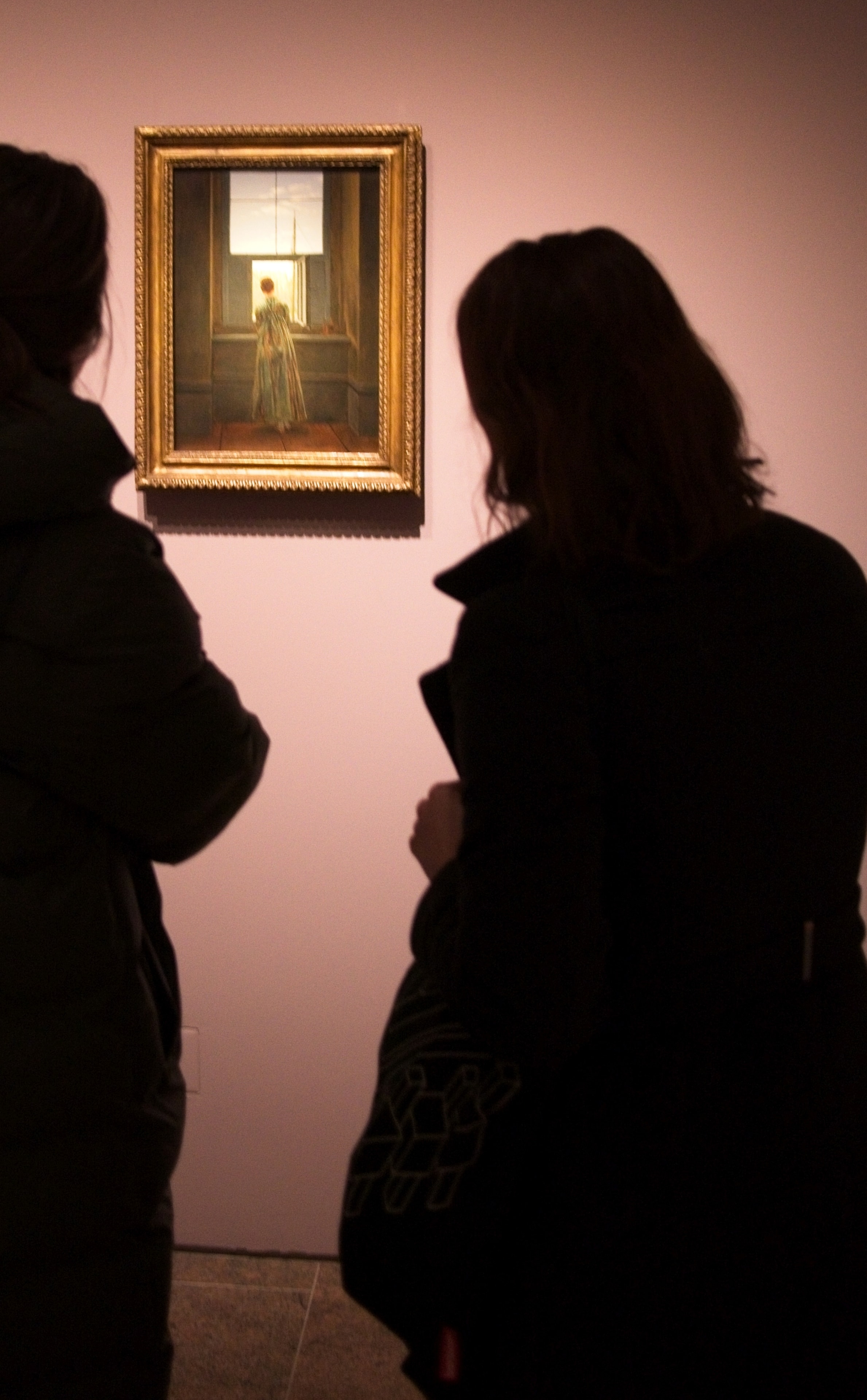 Two silhouetted figures study a candlelit painting of a woman at a window in a dimly lit gallery