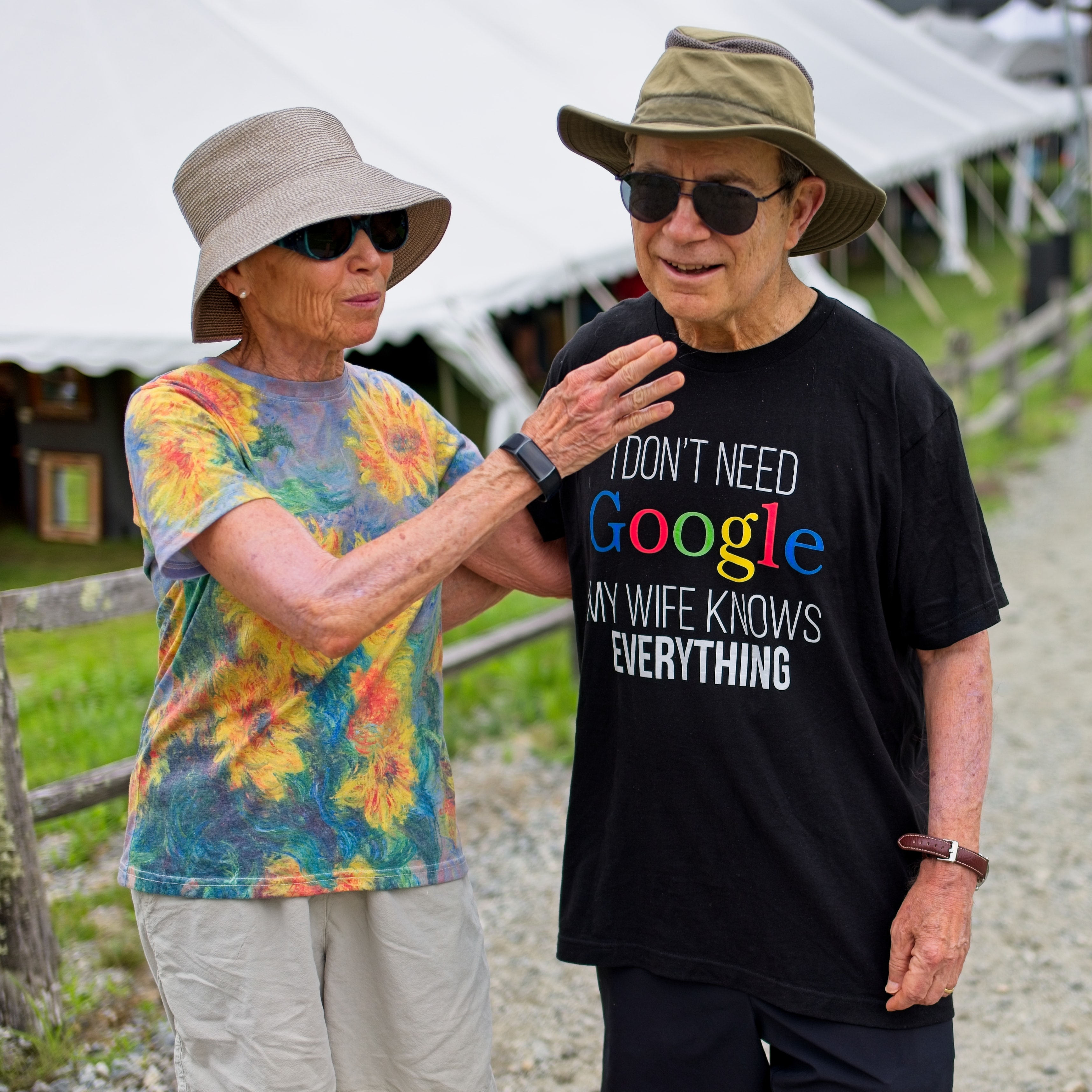 Older couple at an outdoor fair — man wears an 'I Don't Need Google My Wife Knows Everything' t-shirt as his wife gestures at his chest