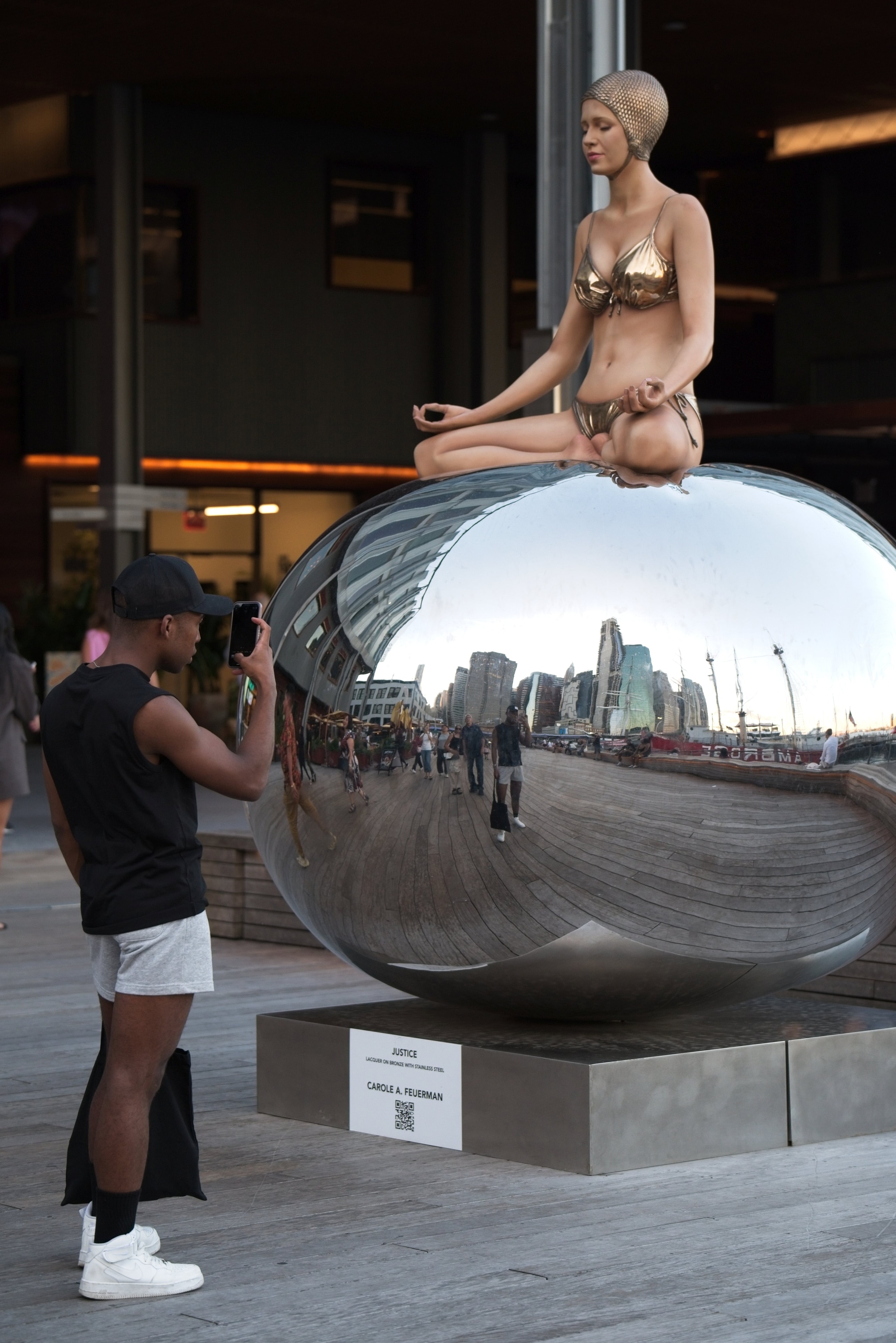 Man photographs the 'Justice' bronze sculpture atop a mirrored sphere at the NYC waterfront, skyline reflected in the orb