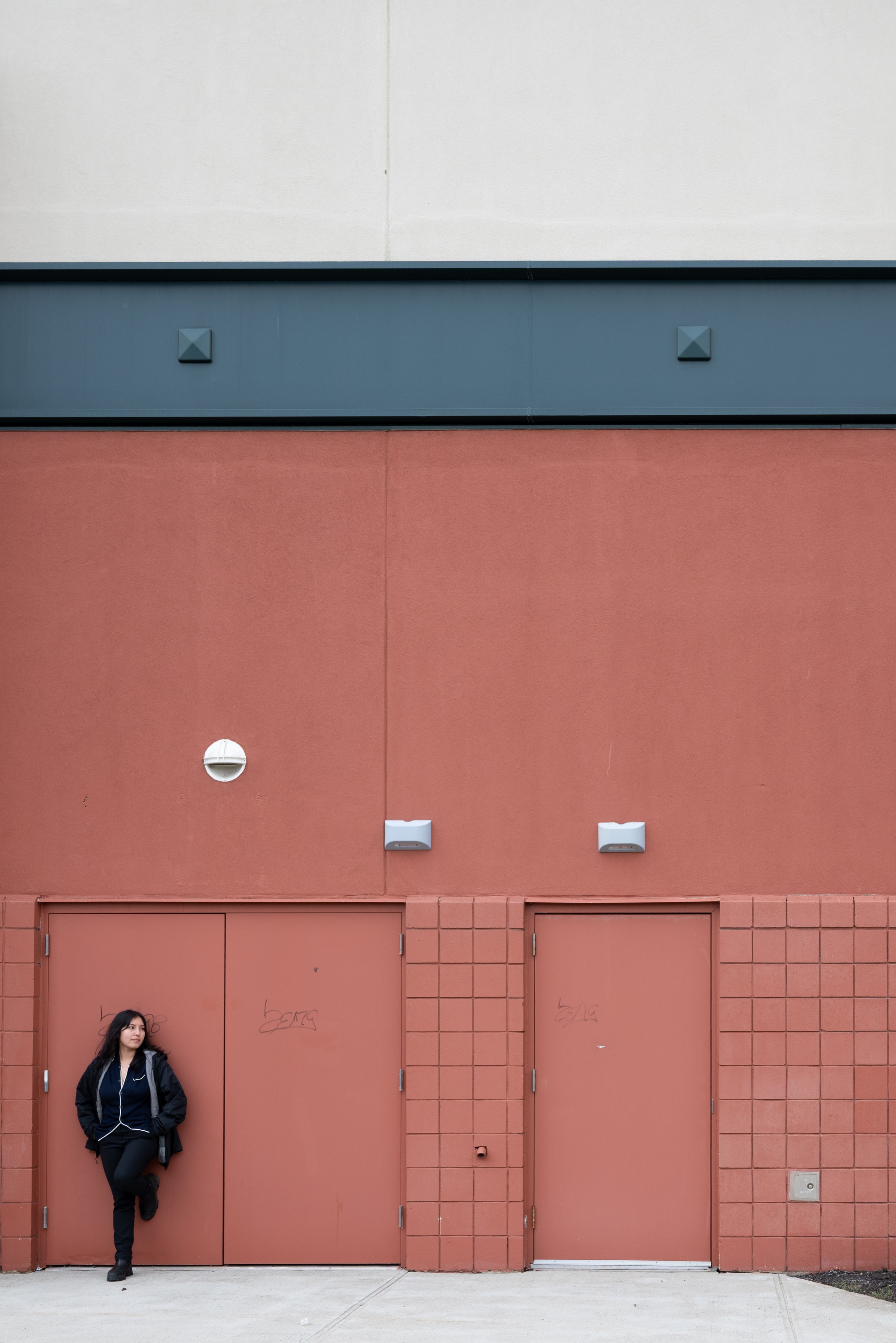 Woman in a black jacket leans against a terracotta-red wall with two metal doors