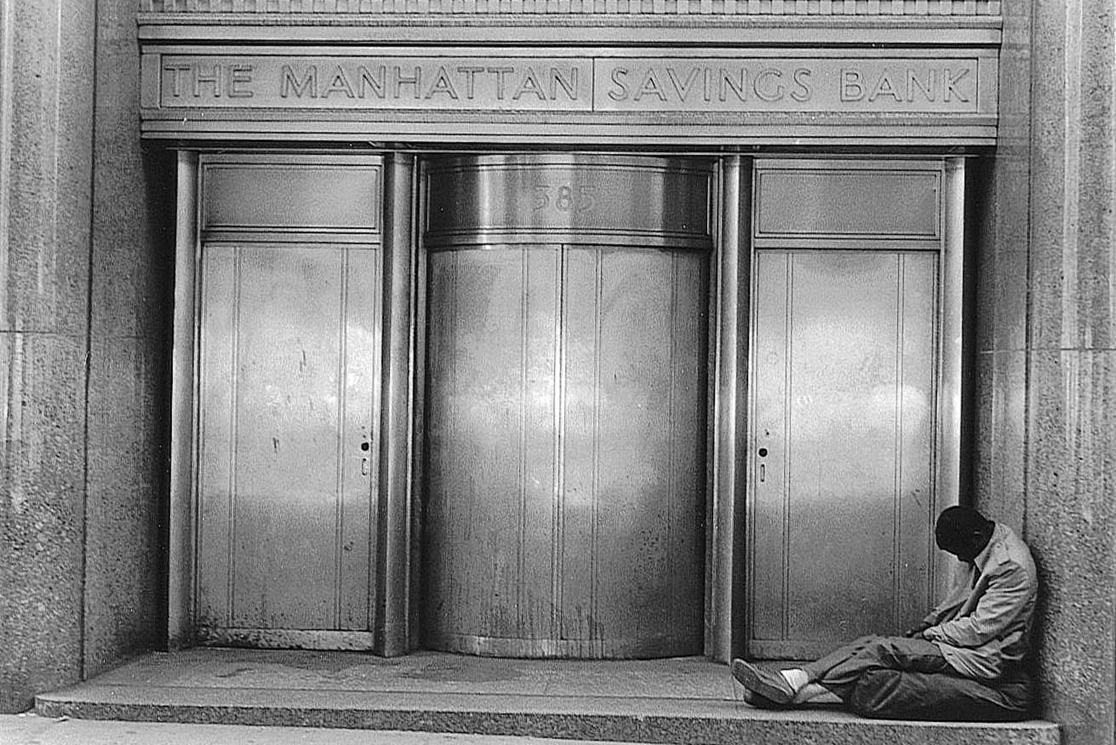 Black and white film photograph of a man slumped against the ornate bronze doors of The Manhattan Savings Bank
