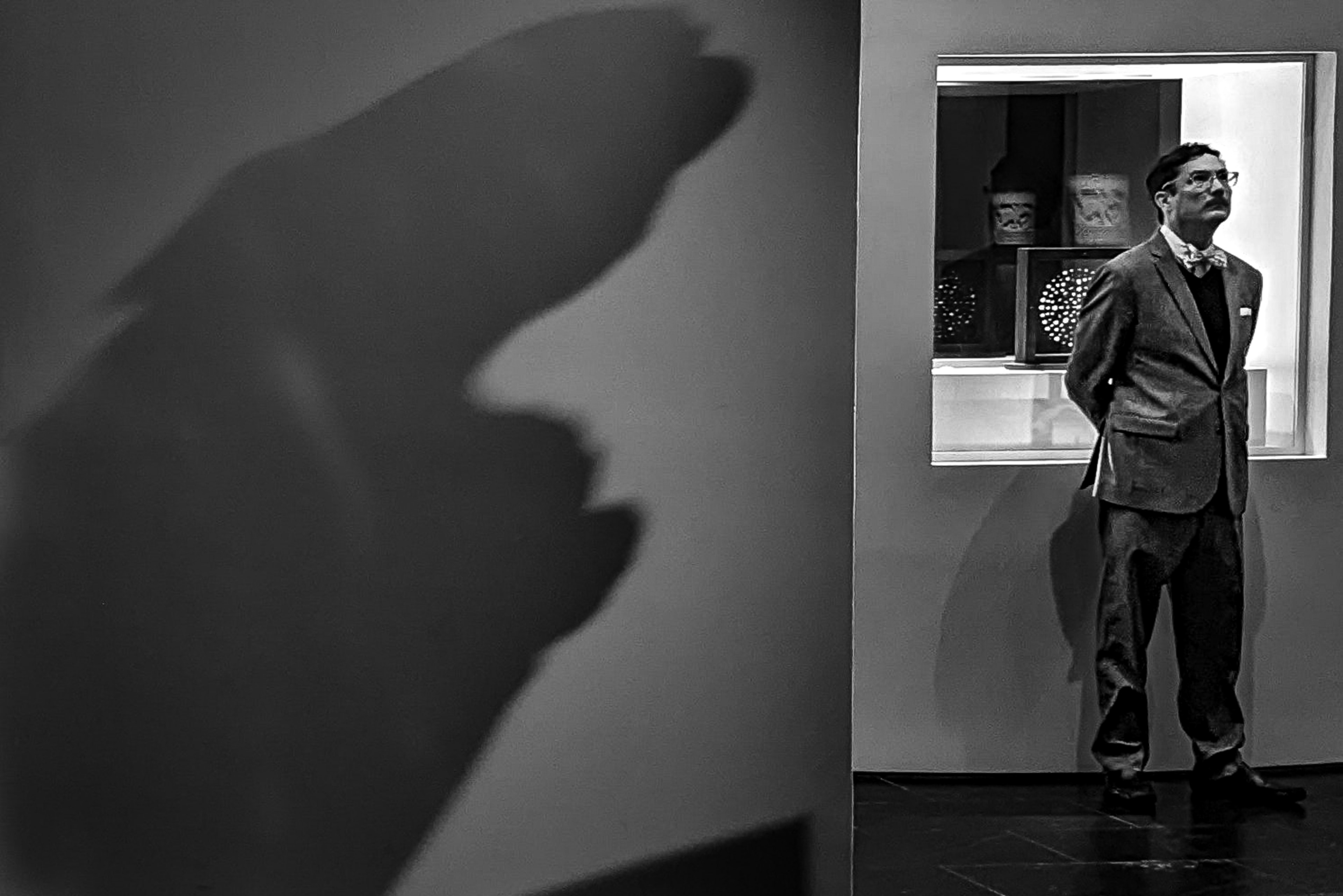 Black and white photograph of a dapper man in a bow tie standing beside a large abstract shadow cast on a museum gallery wall