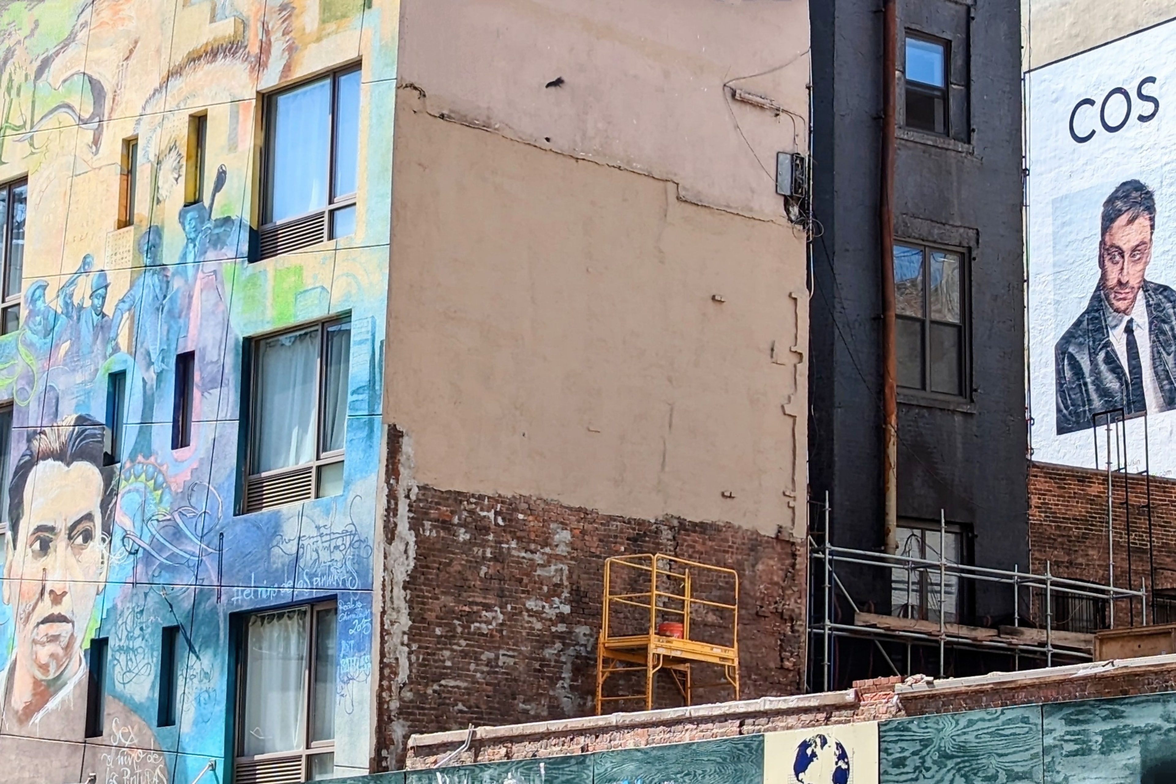 Colorful murals cover a building facade beside a bare concrete wall and scaffolding on a NYC street