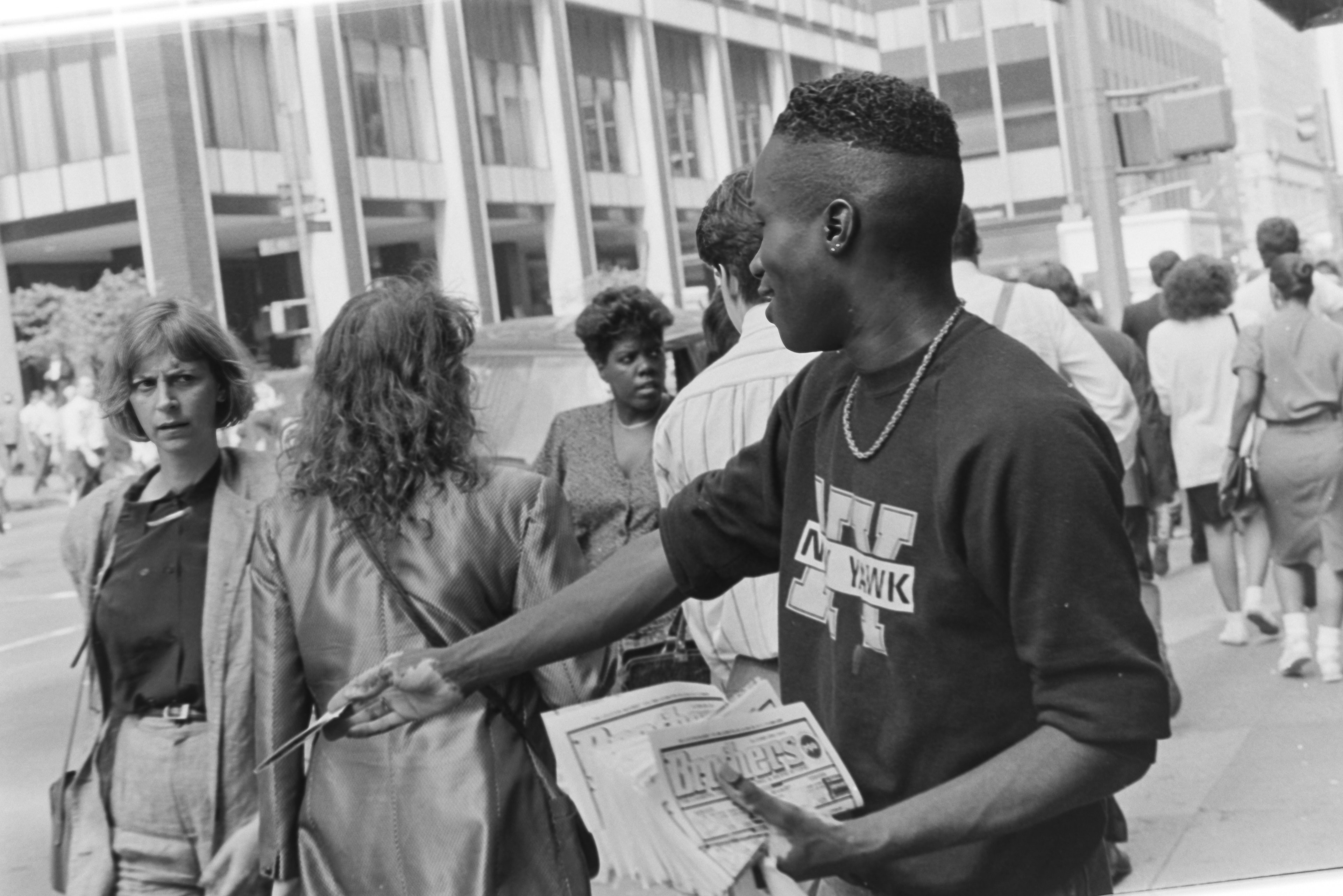 Black and white candid of a man handing a newspaper to a woman on a busy NYC sidewalk