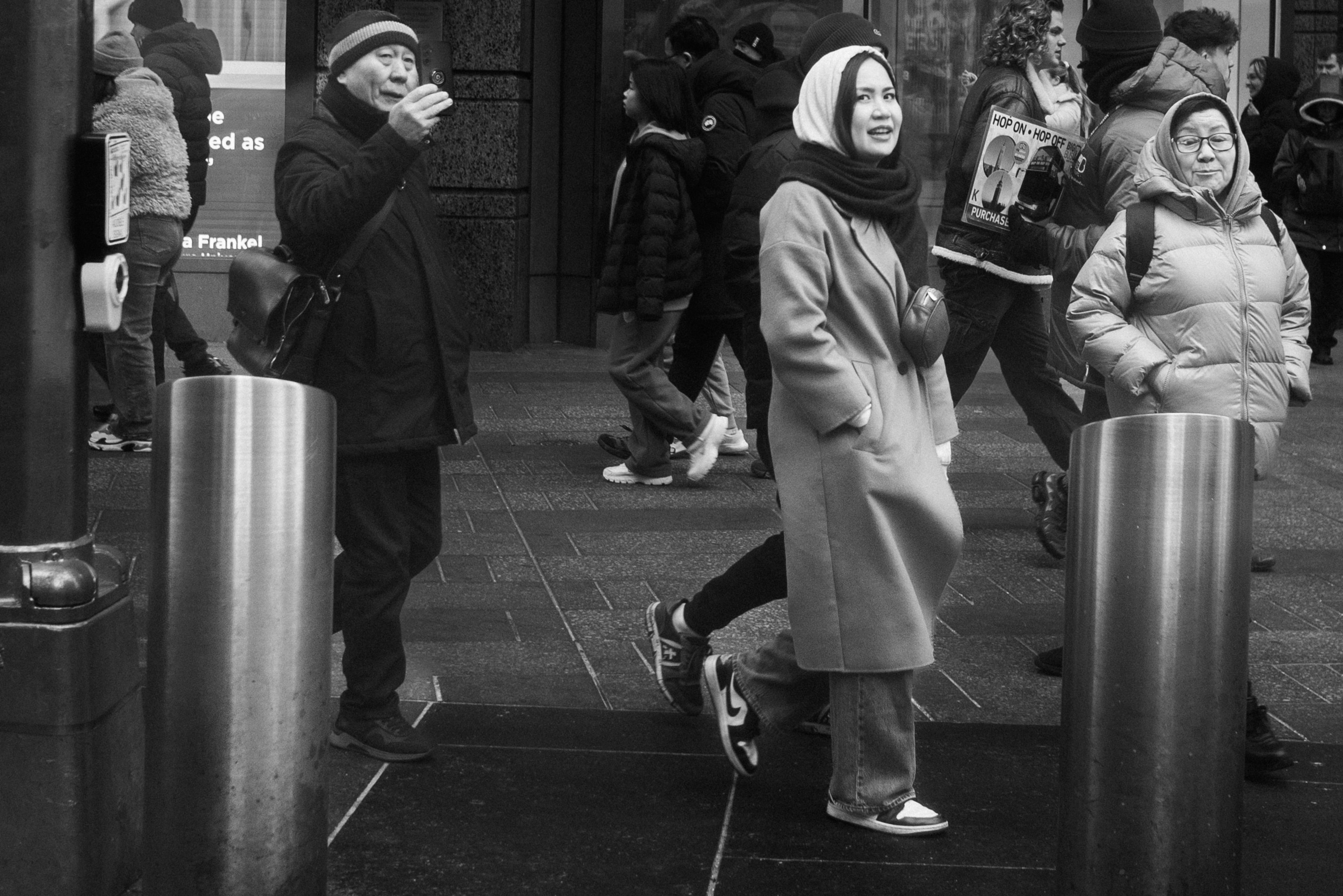 Black and white candid of pedestrians streaming past steel bollards on a busy NYC sidewalk