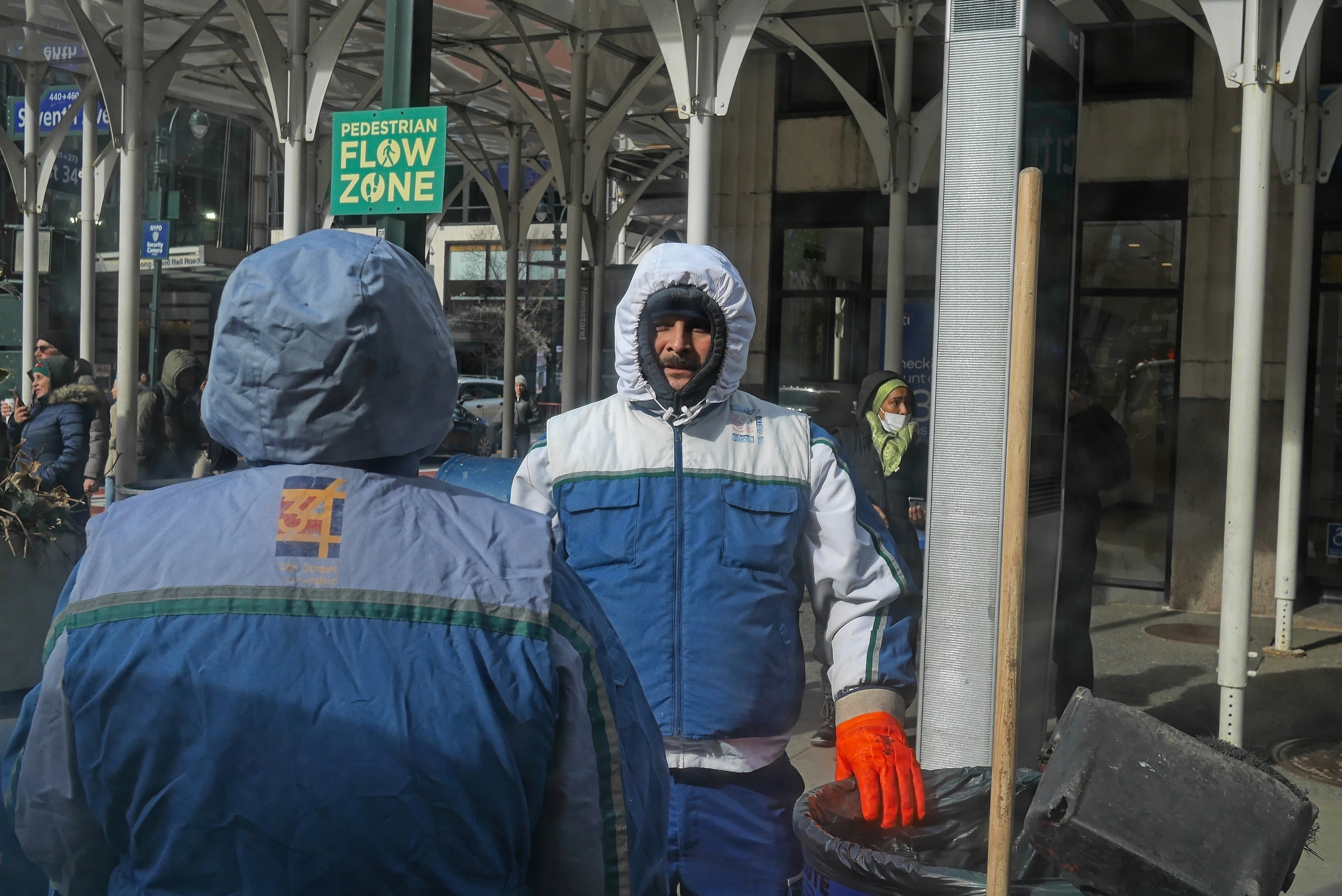 Two sanitation workers in blue uniforms talk beside a Pedestrian Flow Zone sign near Penn Station