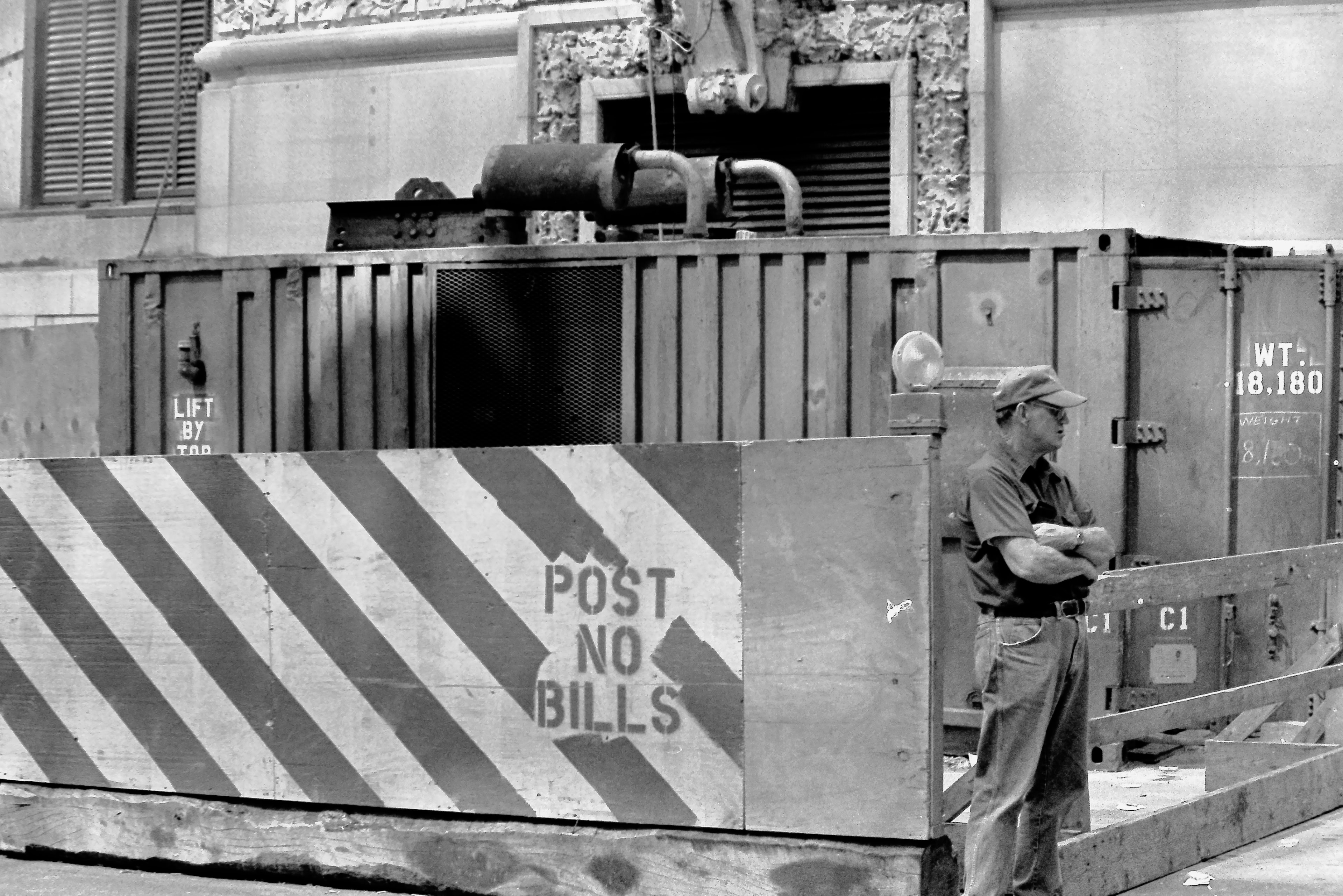 Black and white film photograph of a worker standing beside a Post No Bills construction barrier on a city street