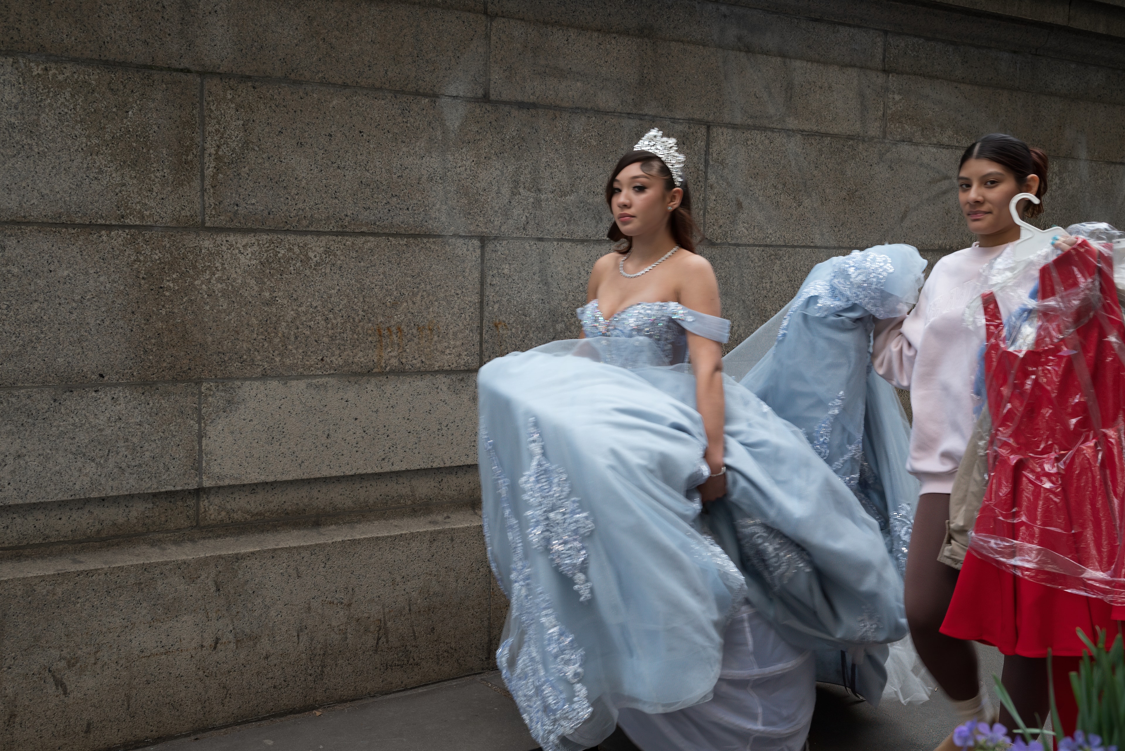 Young woman in a flowing blue quinceañera gown sweeps her skirt against a stone wall while an attendant holds garment bags