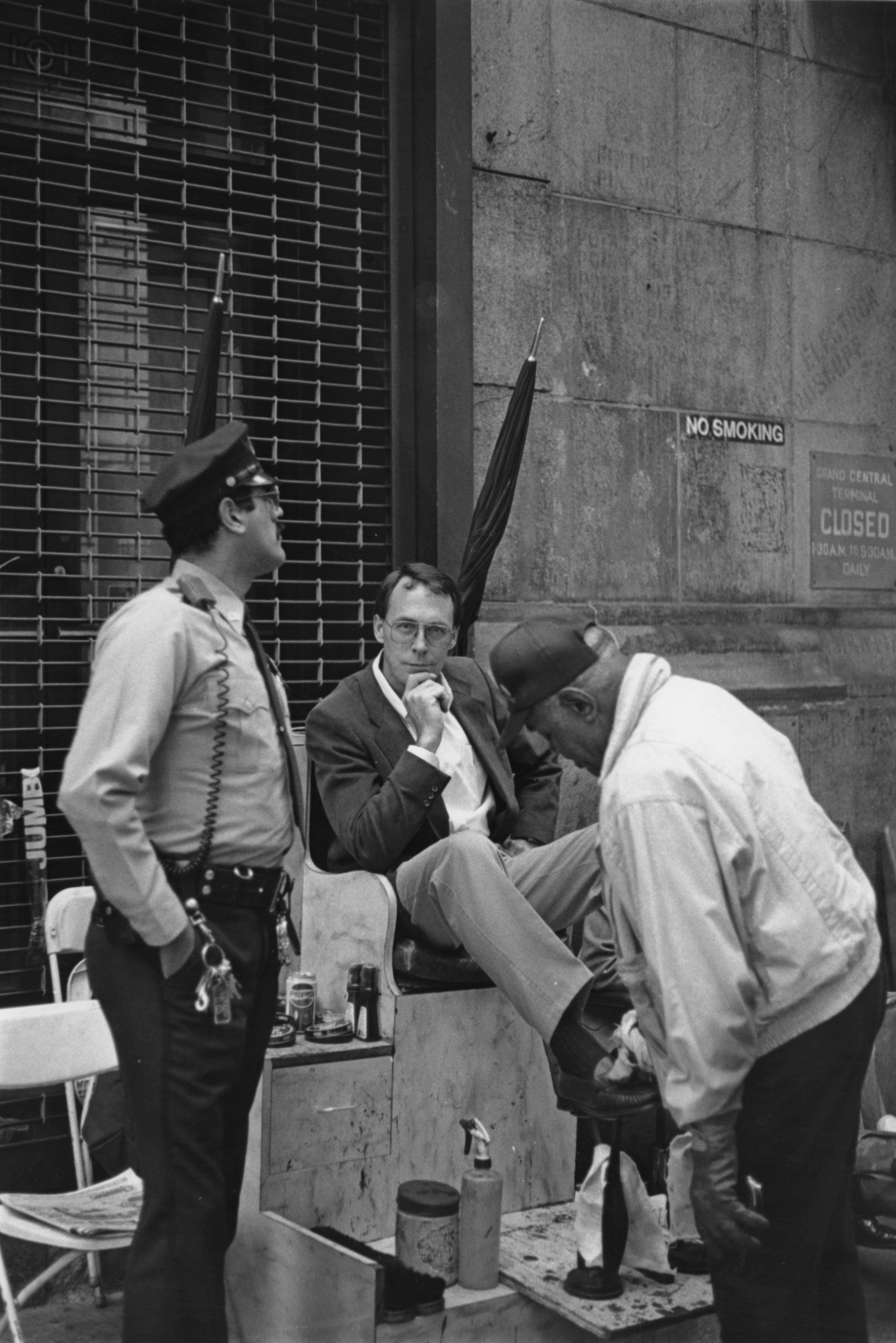 Black and white film photograph of a man getting his shoes shined outside Grand Central Terminal while a security guard looks on