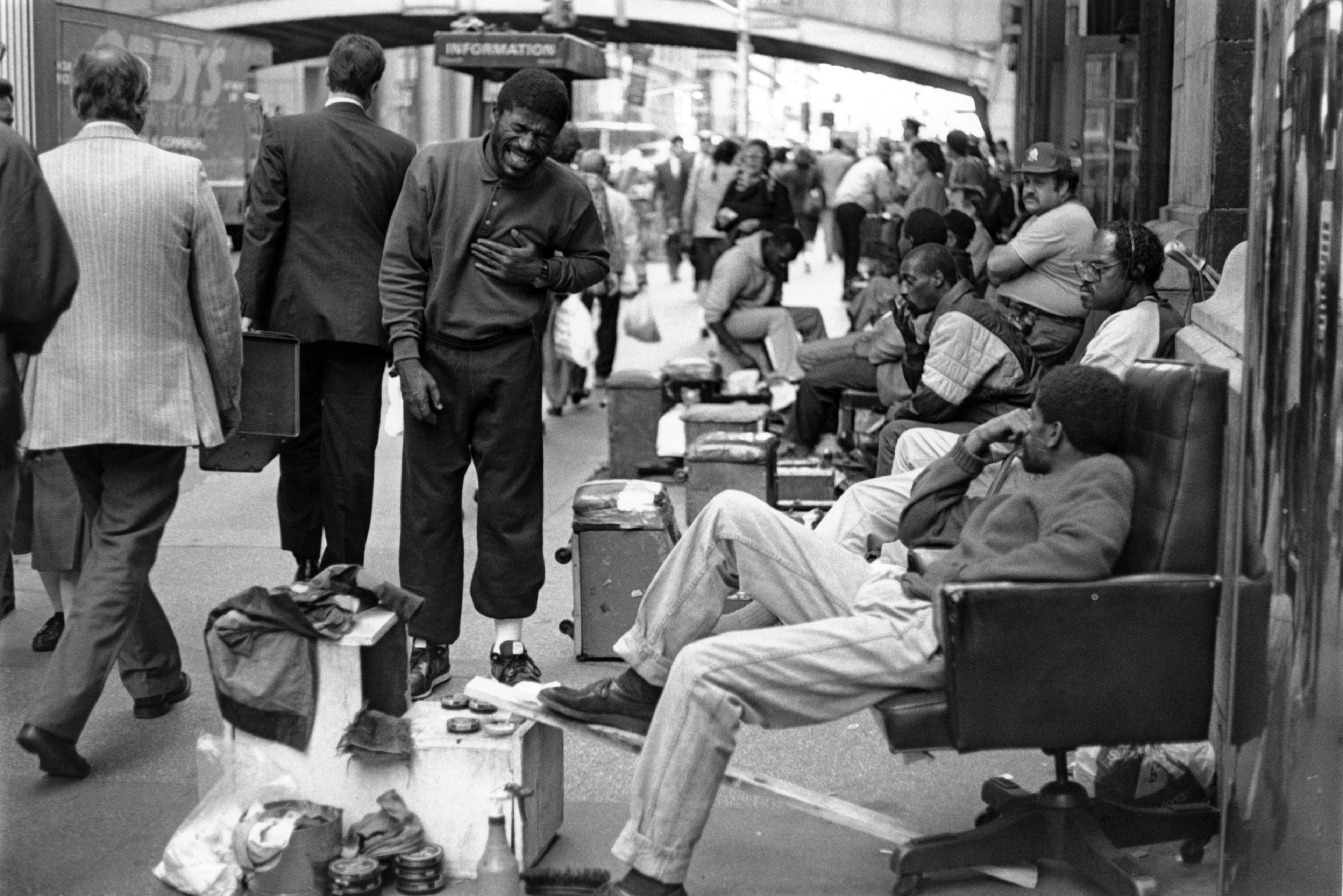 Black and white film photograph of a laughing man among a group of shoeshine workers relaxing on chairs along a busy NYC sidewalk