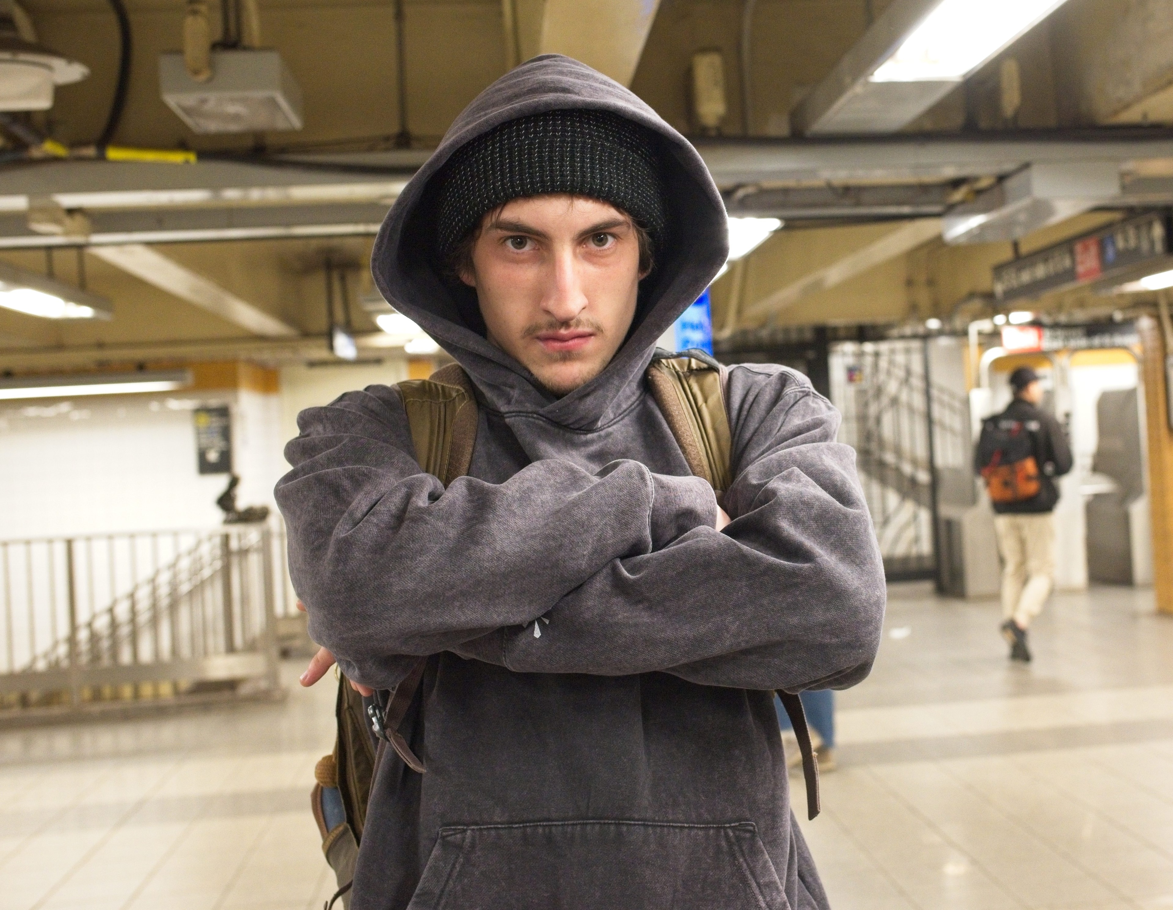 Young man in a hooded sweatshirt stands arms-crossed in a New York City subway station