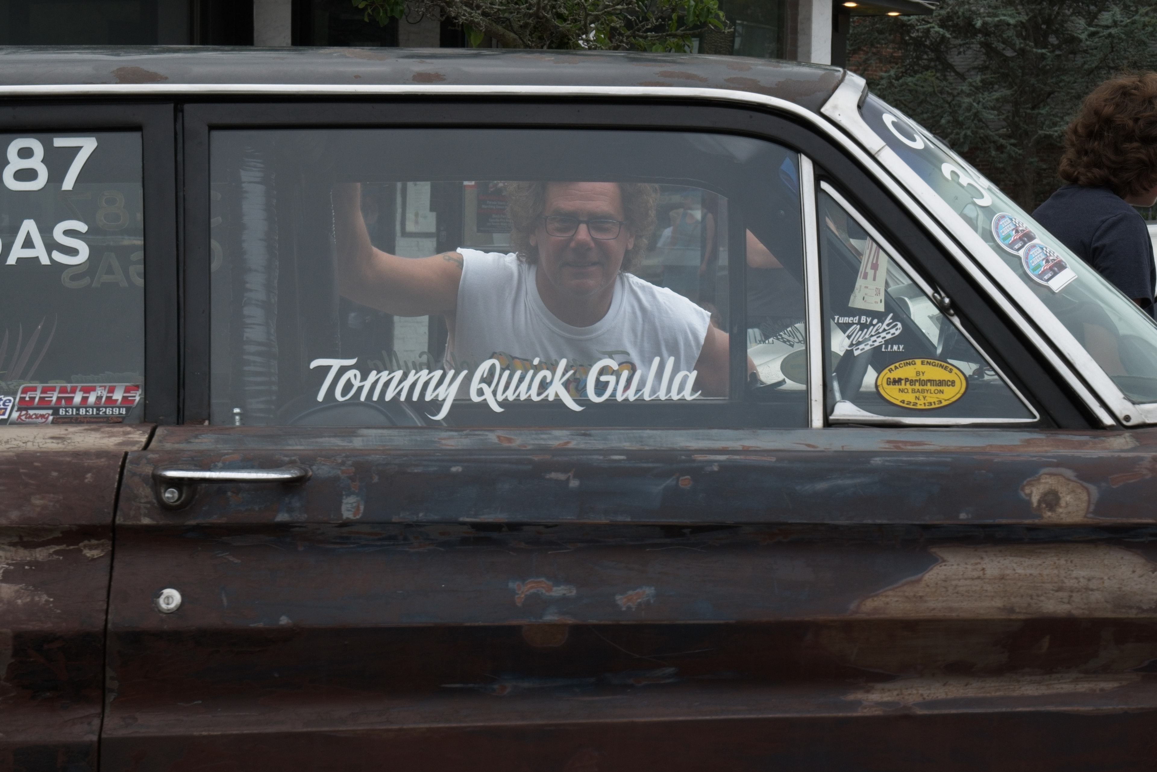Smiling man behind the wheel of a weathered race car with 'Tommy Quick Gulla' lettered on the window