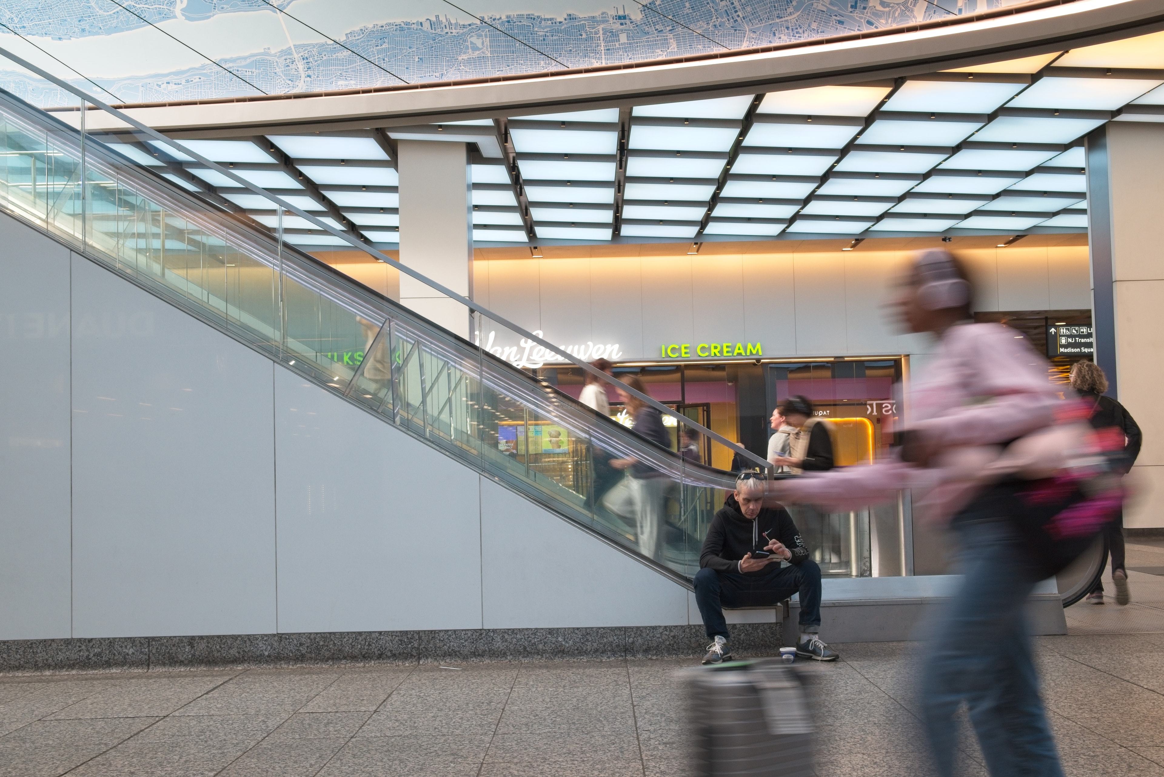 Motion-blurred commuter passes a skateboarder sitting beneath an escalator at a transit hub